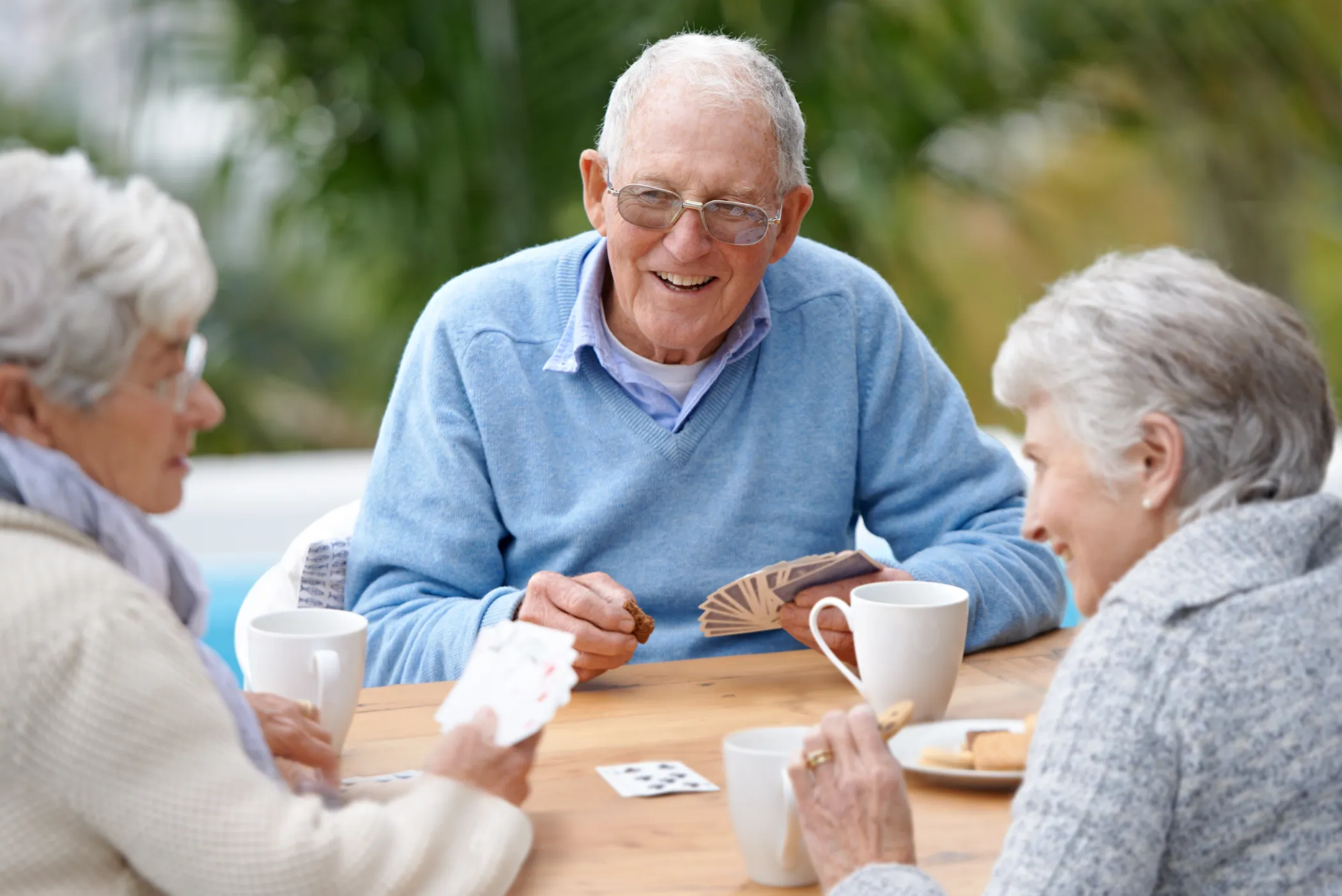 Three elderly people are sitting at a wooden table outdoors, playing cards and enjoying cups of coffee or tea.
