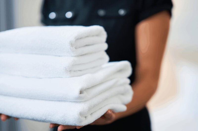 Close-up of a person in a dark uniform holding a stack of four neatly folded, fluffy white towels.