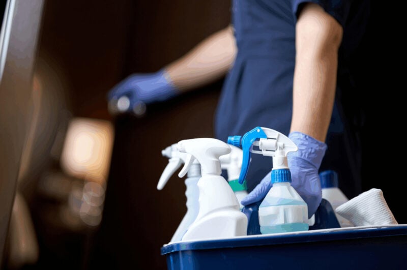 Professional housekeeper wearing blue gloves holding a spray bottle while carrying a caddy of cleaning products.